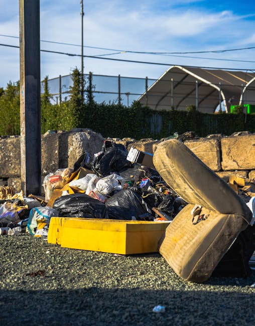 A close-up view of a pile of mixed rubbish accumulated on a gravel surface beside a stone wall. The waste includes several black plastic bags, some partially open revealing scattered paper and plastic waste inside, which appear crumpled and used. There is also a large, beige upholstered sofa armrest or cushion, partially resting on the rubbish and leaning against the stone wall. To the front, a yellow plastic container or tray is positioned among the debris. In the background, a metal fence and some greenery, including shrubs and trees, are visible behind the stone wall. Overhead, a utility pole with wires extends into a blue sky with a few wispy clouds. To the right, a large, semi-circular fabric tent or structure, possibly part of a sports or event facility, stands behind the fence, with bright green signage indicating a sports-related context. The scene is illuminated by natural daylight, emphasizing the textures of the discarded materials and surrounding environment, reflecting a typical example of waste left for private rubbish clearance services such as Rubbish Clearance Highbury, highlighting the need for professional rubbish removal and alternative waste handling solutions.