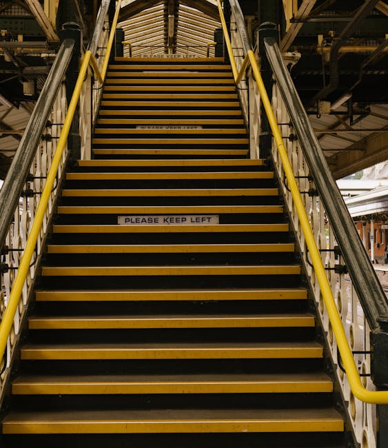 A set of industrial metal stairs with black treads and yellow safety edges, leading upwards in an indoor environment, possibly a warehouse or factory. The stairs have handrails on both sides painted in bright yellow, providing support for users. The background shows structural steel beams, pipes, and other industrial fixtures, indicating a high-bay storage or manufacturing setting. A sign affixed to the middle of the stairs reads 'PLEASE KEEP LEFT,' suggesting a safety or directional instruction. The lighting appears to be artificial, casting a consistent illumination across the scene. The overall environment appears utilitarian with a focus on functionality and safety, aligning with a setting where waste removal or site clearance activities could take place, especially in reference to independent or private disposal methods handled by companies like Rubbish Clearance Highbury.