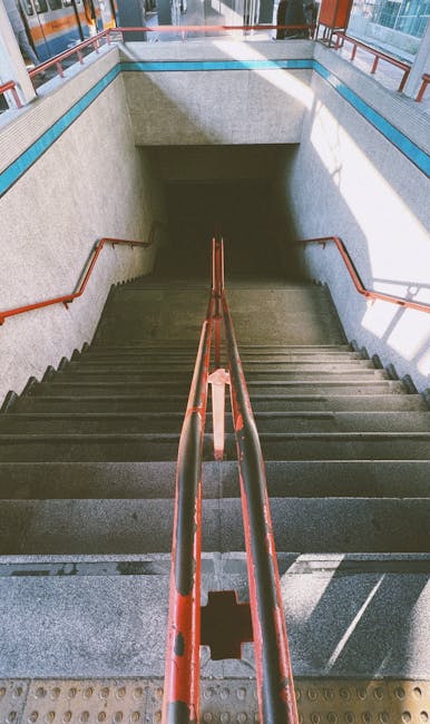 A downward view of a concrete staircase leading to an underground passage or basement, with dark stairs that have textured, slightly worn surfaces. The staircase is flanked by metal handrails painted in red, which are slightly chipped and show signs of wear, running along both sides and converging toward the bottom. The surrounding walls are made of smooth, light-colored concrete, with a blue protective strip running horizontally along the top edge of the wall. At the top of the stairs, there is an open rectangular entrance with a shadowed area beyond, leading to a darker space below. Natural light illuminates the upper part of the scene, casting soft shadows on the stair treads and railings, suggesting a well-lit environment, possibly in a public or commercial building with external access points. The scene captures a clean yet utilitarian setting, typical of an urban infrastructure designed to facilitate access to subterranean levels, aligning with private or independent access solutions, similar to those managed by local waste or rubbish removal services like Rubbish Clearance Highbury, for service-related clearance of underground or basement rubbish storage areas.