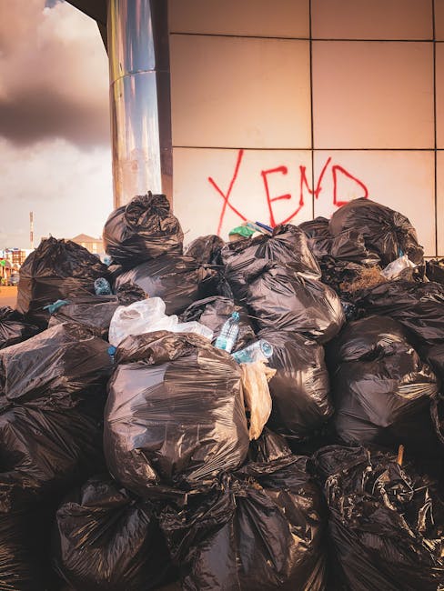 A large pile of black plastic rubbish bags stacked against a modern building with a tiled exterior wall. Some of the bags are torn open, revealing plastic bottles, paper, and other waste materials inside. The bags are unevenly arranged in a dense cluster, with a few white plastic bags and bottles visible among them. In the background, partially obscured by the rubbish, there is graffiti in red spray paint on the wall that reads 'XEND'. The scene appears to be outdoors on a paved surface, possibly adjacent to a commercial or residential building, with dull grey clouds in the sky above. The image suggests a scenario of waste accumulation that could be part of a private rubbish clearance or alternative waste handling service, such as an emergency rubbish removal after eviction, as offered by Rubbish Clearance Highbury.