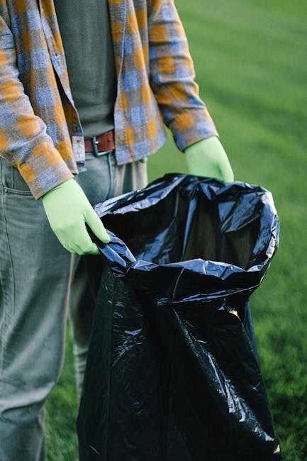 A person wearing a checkered shirt in shades of grey, blue, and brown, along with a belt and grey trousers, is holding an open black rubbish bag lined with a silver interior on a grassy outdoor area. The individual is wearing light green gloves, indicating they are engaged in waste collection or rubbish removal. The background shows a blurred expanse of green grass, suggesting a park or garden setting. The person's torso and arms are visible, but their face is not included. The scene depicts a private or independent rubbish clearance activity, possibly related to environmentally friendly waste handling or garden tidy-up, reflective of services offered by companies like Rubbish Clearance Highbury for on-site rubbish removal and disposal. The lighting is natural, and the focus is on the process of preparing the rubbish bag for collection, emphasizing careful handling of waste materials in a professional manner.