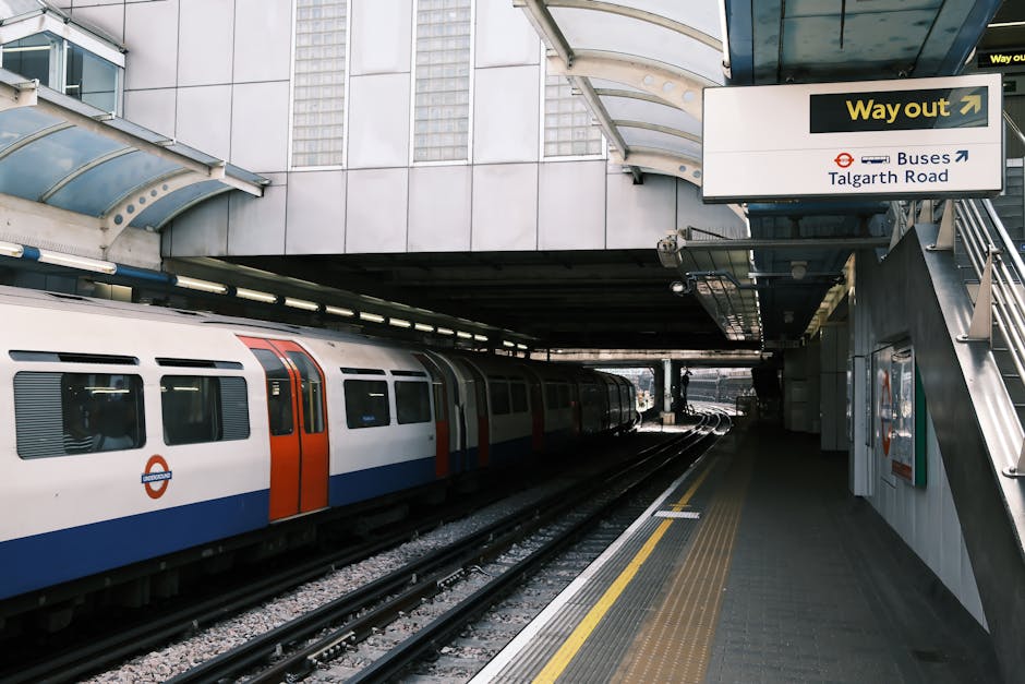 A London Underground train with a white body, orange and blue stripe accents, and the roundel logo on the door, parked on tracks at an outdoor station platform. The platform has a textured, non-slip surface with yellow tactile paving along the edge for safety. Overhead, there is a partially covered structure with a curved, translucent roof supported by metal beams, providing shelter for passengers. In the background, a sign indicates an exit heading towards Talgarth Road and bus connections, with an arrow pointing right. The station is adjacent to modern multi-story buildings with glass and metal facades, and the environment appears urban and well-lit with natural daylight. This scene emphasizes an alternative to private waste disposal, fitting with the context of rubbish removal services by professionals, such as Rubbish Clearance Highbury, although no rubbish or removal activity is visible in the image.