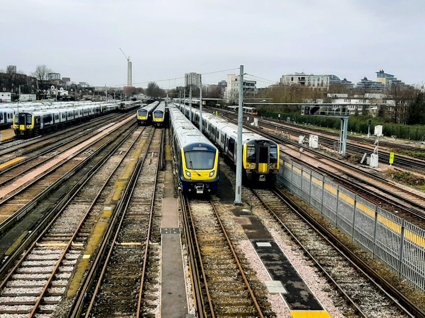 A railway yard with multiple train tracks running parallel across the image, featuring modern yellow and gray passenger trains parked on several lines. The foreground shows two trains side by side, one facing directly toward the viewer and the other slightly angled, with sleek exteriors and tinted windows. The tracks are made of steel rails mounted on wooden sleepers and gravel beds, with some tracks having additional switches and points. To the right, there is a tall metal safety fence topped with yellow safety markings, separating the trains from the surrounding area. In the background, various industrial and commercial buildings are visible, including mid-rise structures, some with windows, and a few taller high-rise buildings further away. The sky overhead is overcast with gray clouds, creating diffuse lighting across the scene. The entire setting appears to be a busy rail depot or storage yard, which could be associated with independent or private yet regulated railway operations, relevant to alternatives to public transportation services.
