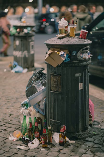 A black metal public trash bin situated on a cobblestone sidewalk with a domed lid. The lid is cluttered with various discarded items including paper cups, plastic cups, and food wrappers. Surrounding the base of the bin on the ground are multiple empty glass bottles, mostly beer bottles with red and green labels, as well as crumpled paper and plastic waste. A black garbage bag is tied to the side of the bin, and a cardboard box is leaning against it, partially filled with additional rubbish. In the background, blurred figures of pedestrians and parked cars are visible, with one dark-colored vehicle parked right next to the bin. The scene suggests a busy urban area with litter accumulation near a street. Rubbish Clearance Highbury handles waste collection and disposal, which includes managing similar informal rubbish accumulations in public spaces.