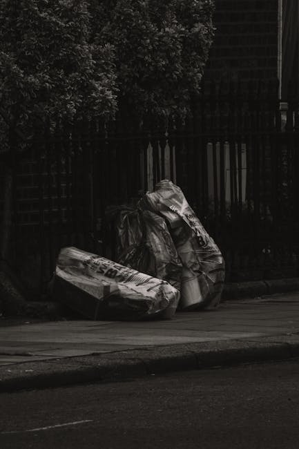 Two large black plastic garbage bags filled with waste material are placed on a street pavement near a black metal fence, with some lightweight white plastic bags visibly protruding from the top of one of the bags. The bags appear to contain mixed refuse, possibly consisting of household or garden waste. Behind the fence, dense foliage from a bush or tree is partially visible, and the scene is illuminated by what appears to be natural daylight, creating a contrast between the dark fence and the lighter bags. The setting suggests a private property or an area where rubbish clearance services, such as those provided by Rubbish Clearance Highbury, might collect bulky waste or excess rubbish for disposal, supporting alternative waste handling outside local authority collection schemes.