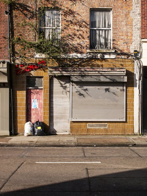 The image shows the front of a small commercial building on a street in an urban area. The lower façade is covered with yellow-brown ceramic tiles, with a large metal roller shutter door in a closed position, painted in light grey. To the left of the shutter is a narrow, metal pedestrian door also in grey, with a small vent below it. In front of the building, near the curb, there are three black rubbish bags and one transparent bag filled with discarded waste, placed against the wall. Above, the building’s upper section reveals two adjacent rectangular windows with white frames, partially covered by curtains inside. The exterior brickwork of the upper story shows red bricks with some weathering and visible mortar lines, and a small leafy green plant extends from the left side, casting shadows onto the brick surface. The street surface is asphalt with white painted lines, indicating a parking or loading zone, and the overall atmosphere suggests a typical urban setting where private waste collection by services like Rubbish Clearance Highbury may be arranged for such waste accumulation.